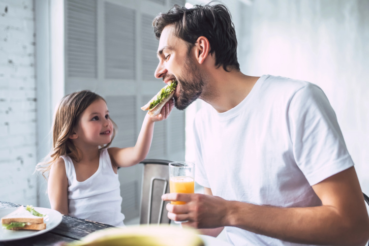 daughter with her father sitting at the table. Daughter is feeding her dad a sandwhich.